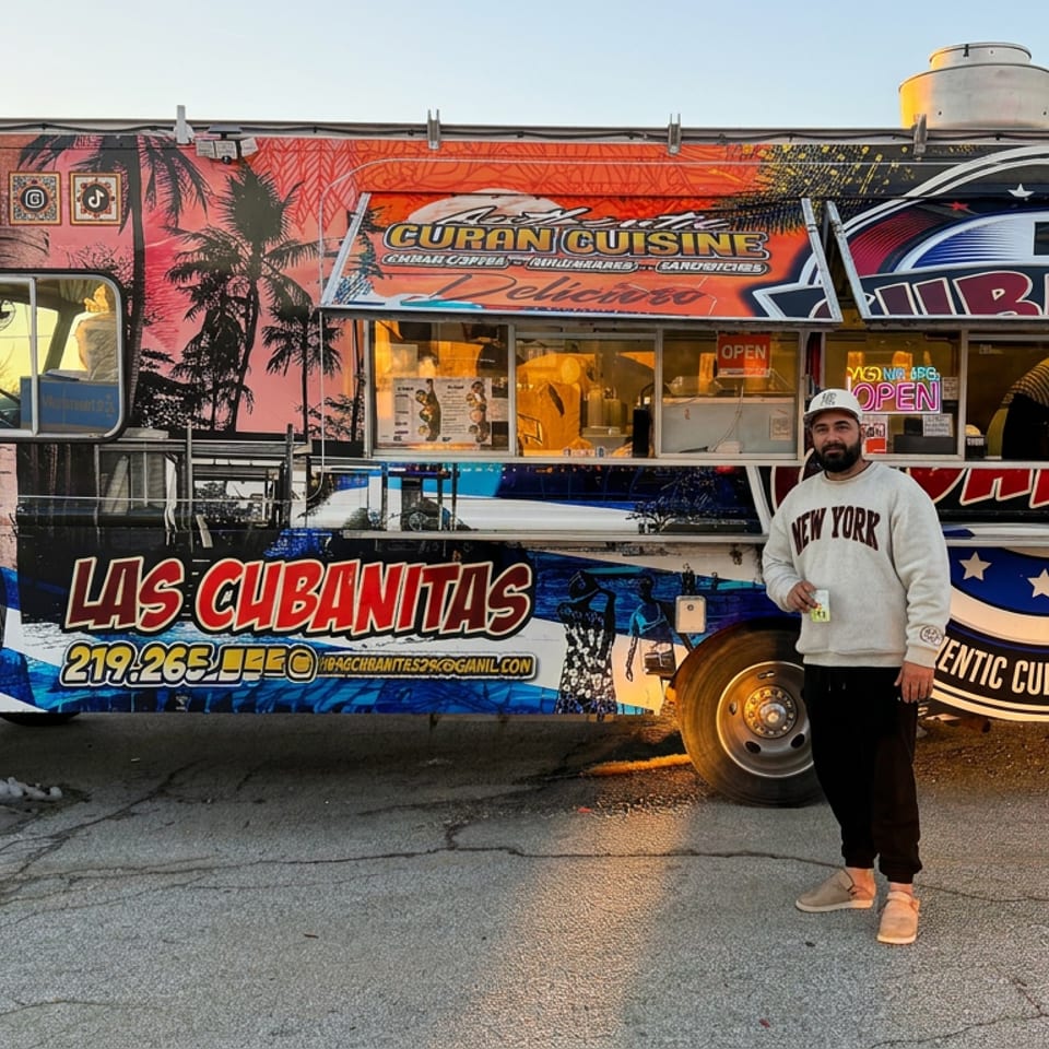 Man stands beside the colorful Las Cubanitas food truck, advertising Cuban cuisine and catering on a snowy street.